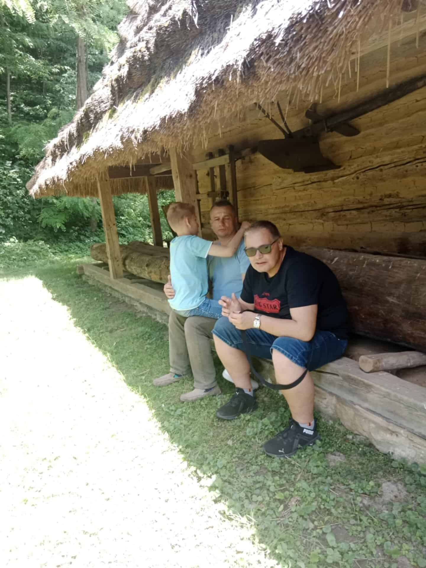 Trzy osoby siedzą na drewnianej ławce pod dachem krytym strzechą; jeden dorosły trzyma dziecko, podczas gdy drugi dorosły siedzi obok nich w słonecznej scenerii na zewnątrz.