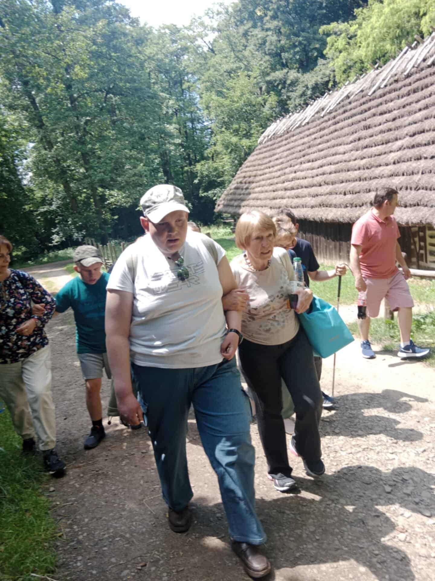 Grupa ludzi idzie razem polną ścieżką obok budynku krytego strzechą w słoneczny dzień.