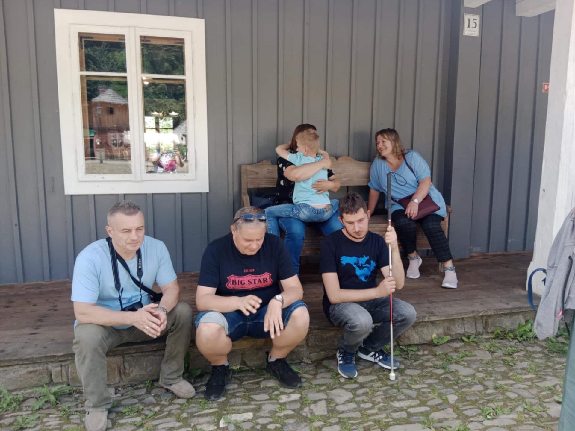 Pięć osób siedzących na drewnianej werandzie; dwie przytulają się, jedna trzyma białą laskę, a dwie inne siedzą cicho. Okno i drzwi są widoczne na drewnianej ścianie za nimi.