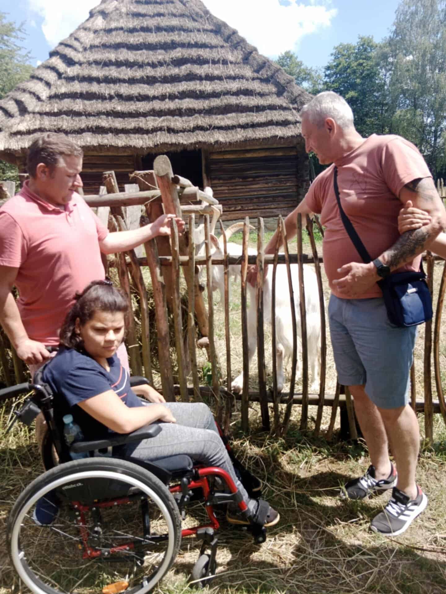 Młoda dziewczyna na wózku inwalidzkim i dwaj mężczyźni stoją przy drewnianym płocie, głaszcząc kozę w pobliżu krytej strzechą konstrukcji na zewnątrz.