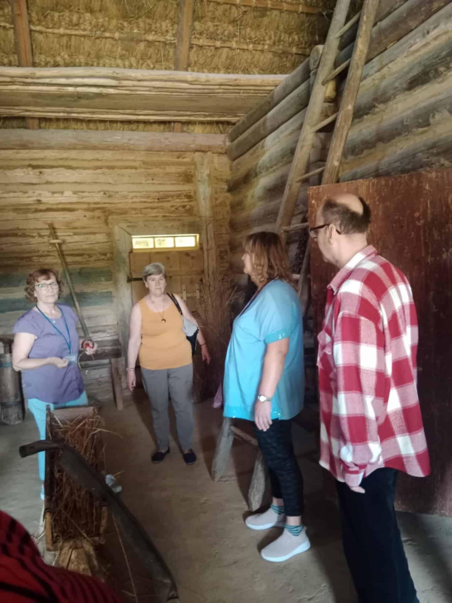 Cztery dorosłe osoby stoją i rozmawiają wewnątrz rustykalnej drewnianej chaty ze ścianami z bali, dachem krytym strzechą i narzędziami rolniczymi widocznymi w tle.