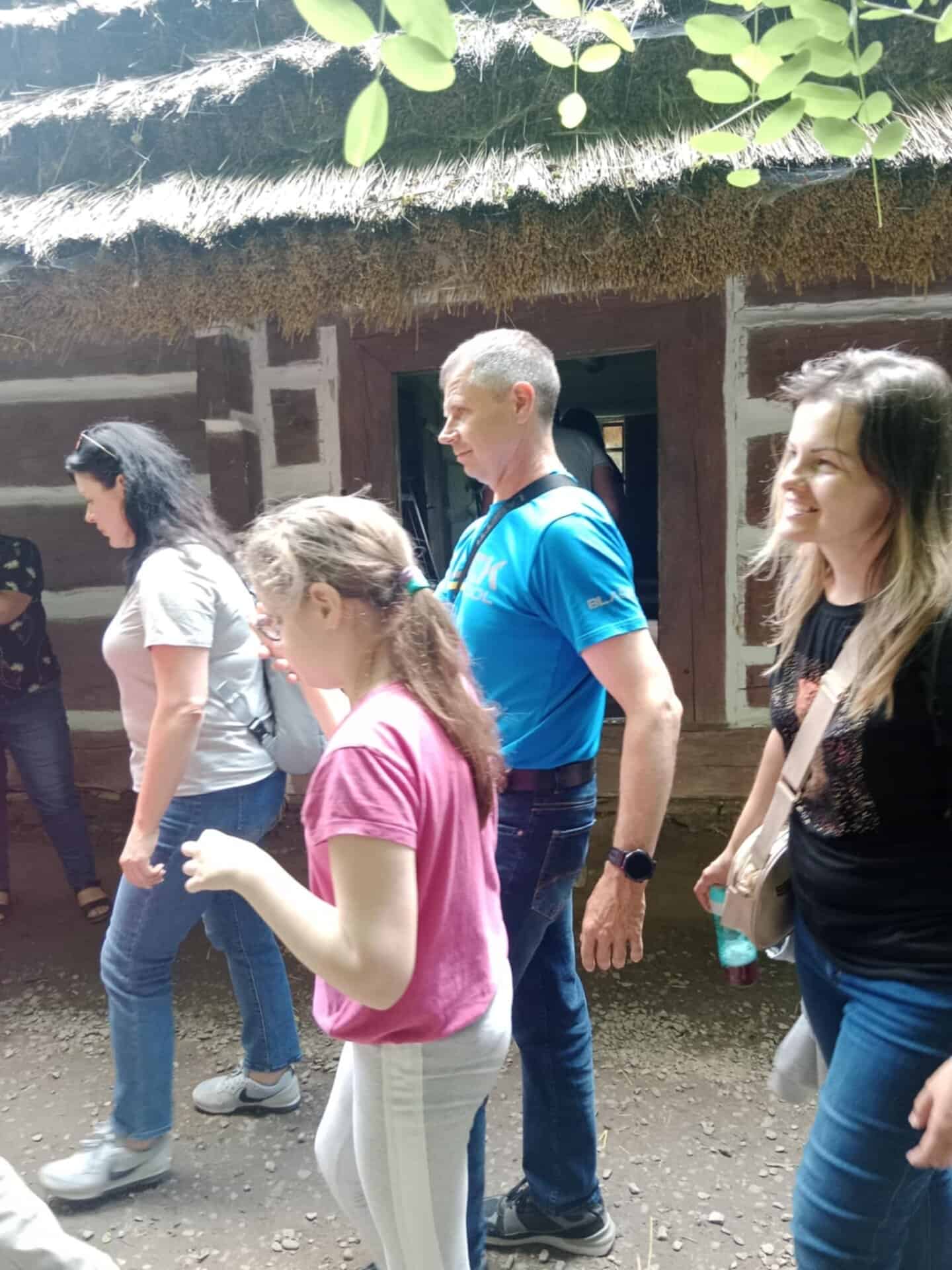 Grupa ludzi przechodzi obok rustykalnego drewnianego budynku z dachem krytym strzechą w słoneczny dzień.