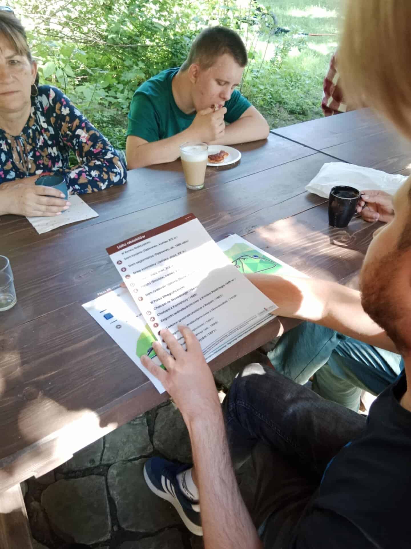 Kilka osób siedzi przy drewnianym stole na świeżym powietrzu; jedna osoba czyta wydrukowane menu, podczas gdy inna ma przed sobą napój i ciasto.