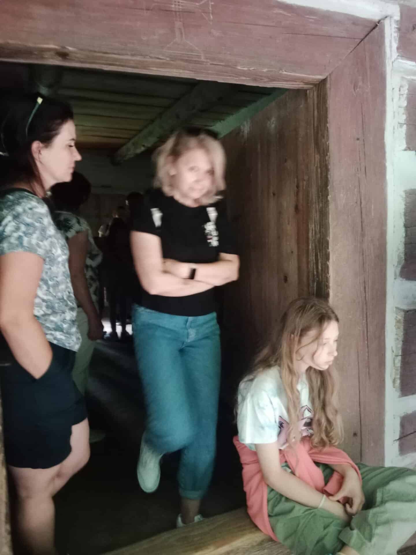 Wewnątrz rustykalnej drewnianej konstrukcji widać trzy osoby; jedna kobieta stoi, druga przechodzi przez drzwi, a dziewczyna z długimi włosami siedzi w drzwiach, patrząc w dół.