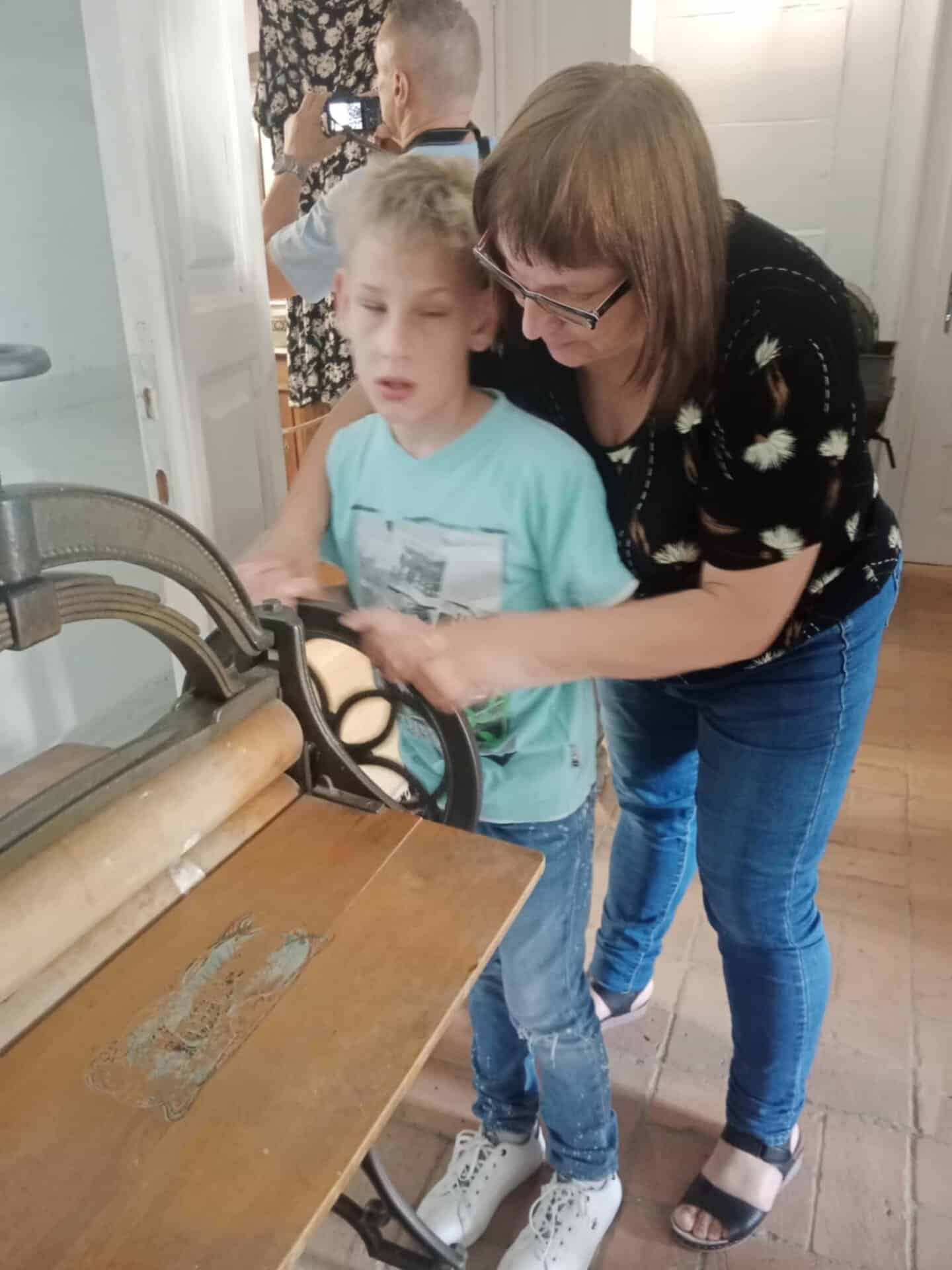 Kobieta pomaga młodemu chłopcu obsługiwać zabytkową maszynę w pokoju, podczas gdy inna osoba robi zdjęcie w tle.