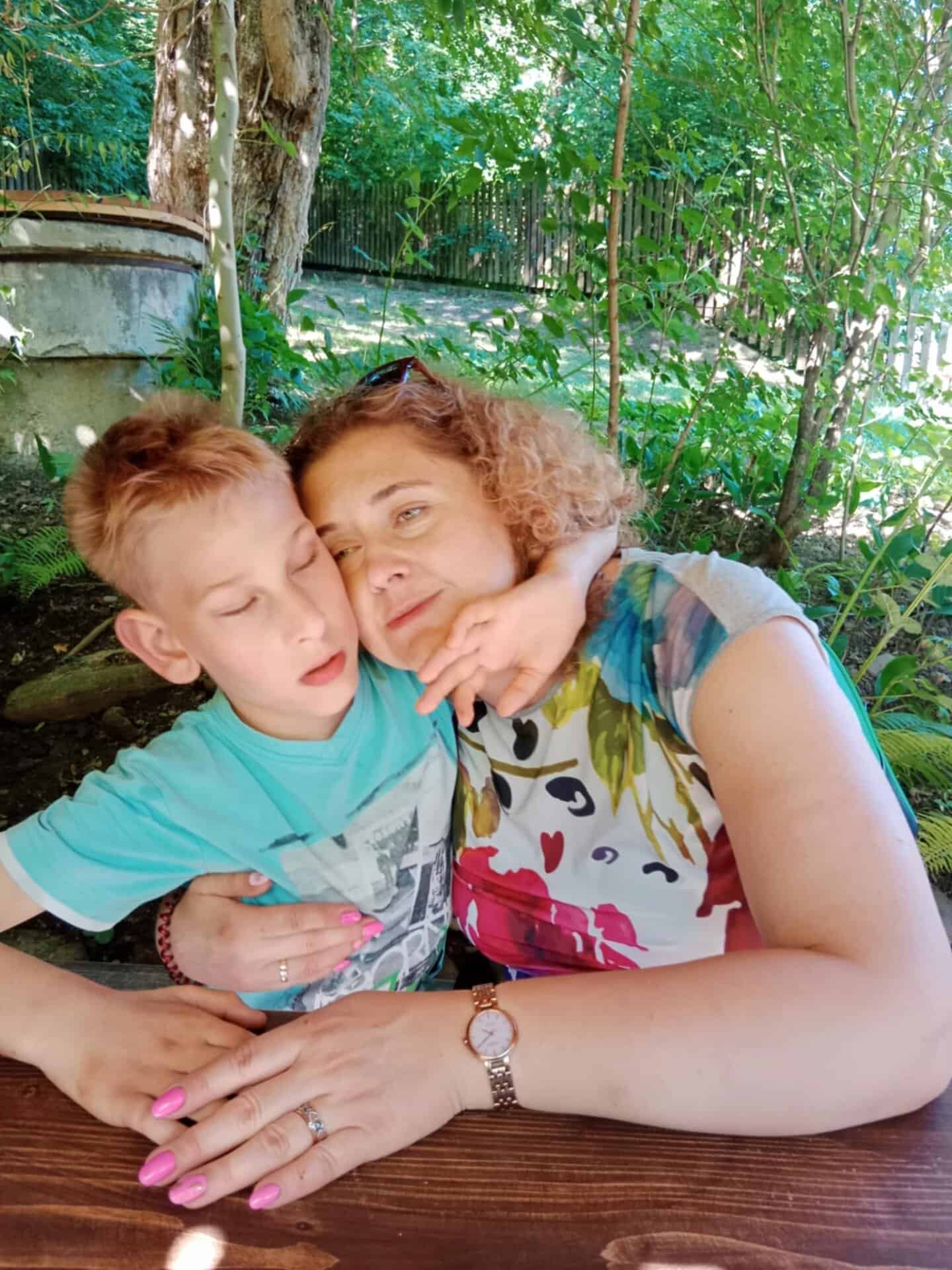 Kobieta z kręconymi włosami przytula małego chłopca przy drewnianym stole na świeżym powietrzu, otoczonym zielenią i drzewami w tle.