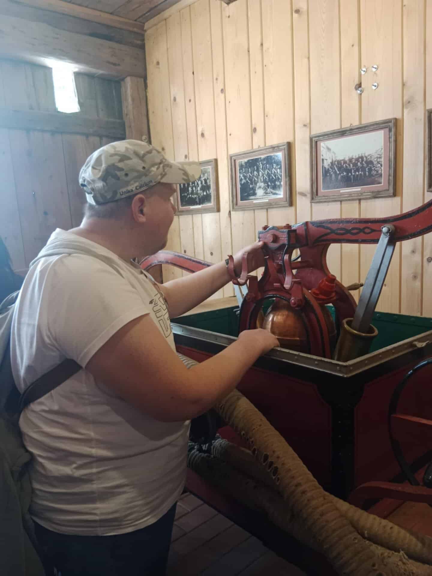 Osoba w czapce i białej koszuli ogląda starą pompę strażacką wystawioną w drewnianym pomieszczeniu z oprawionymi zdjęciami na ścianie.
