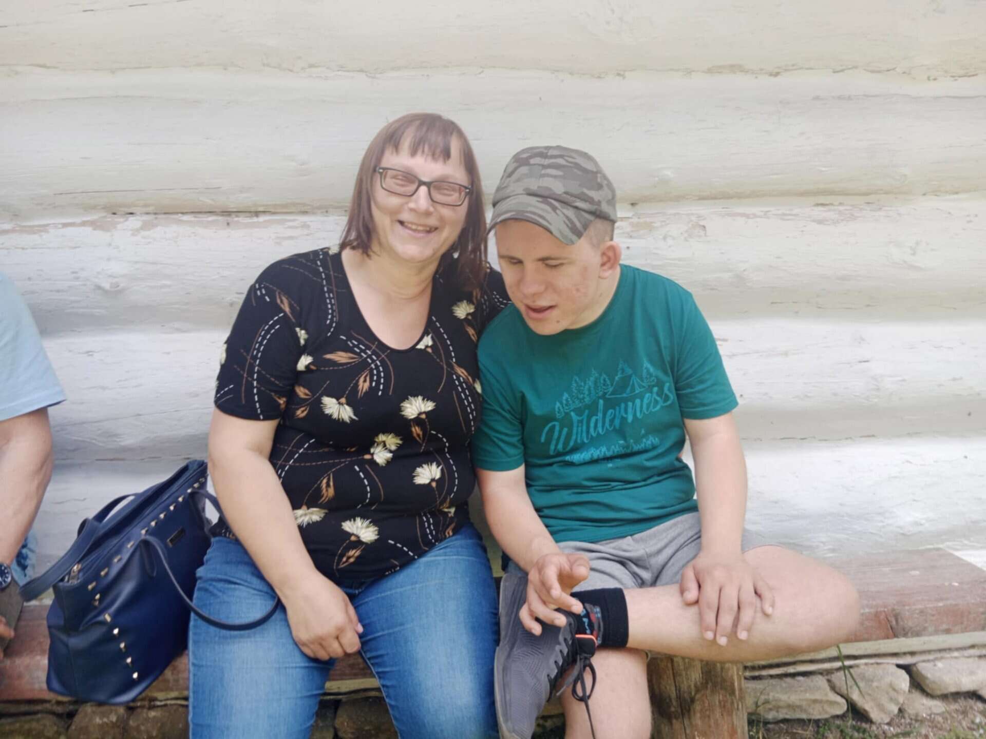 Kobieta i młody mężczyzna siedzą na drewnianej ławce na zewnątrz, uśmiechając się do białej ściany z bali. Kobieta trzyma na kolanach niebieską torebkę, a młody mężczyzna ma na sobie zieloną koszulę i czapkę kamuflażową.