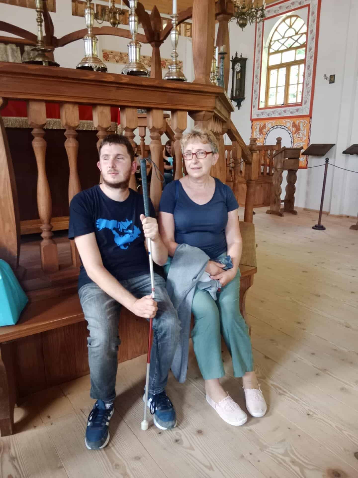 Młody mężczyzna trzymający białą laskę i starsza kobieta siedzą razem na drewnianych schodach wewnątrz jasnego, ozdobnie zdobionego pokoju z dużymi oknami.