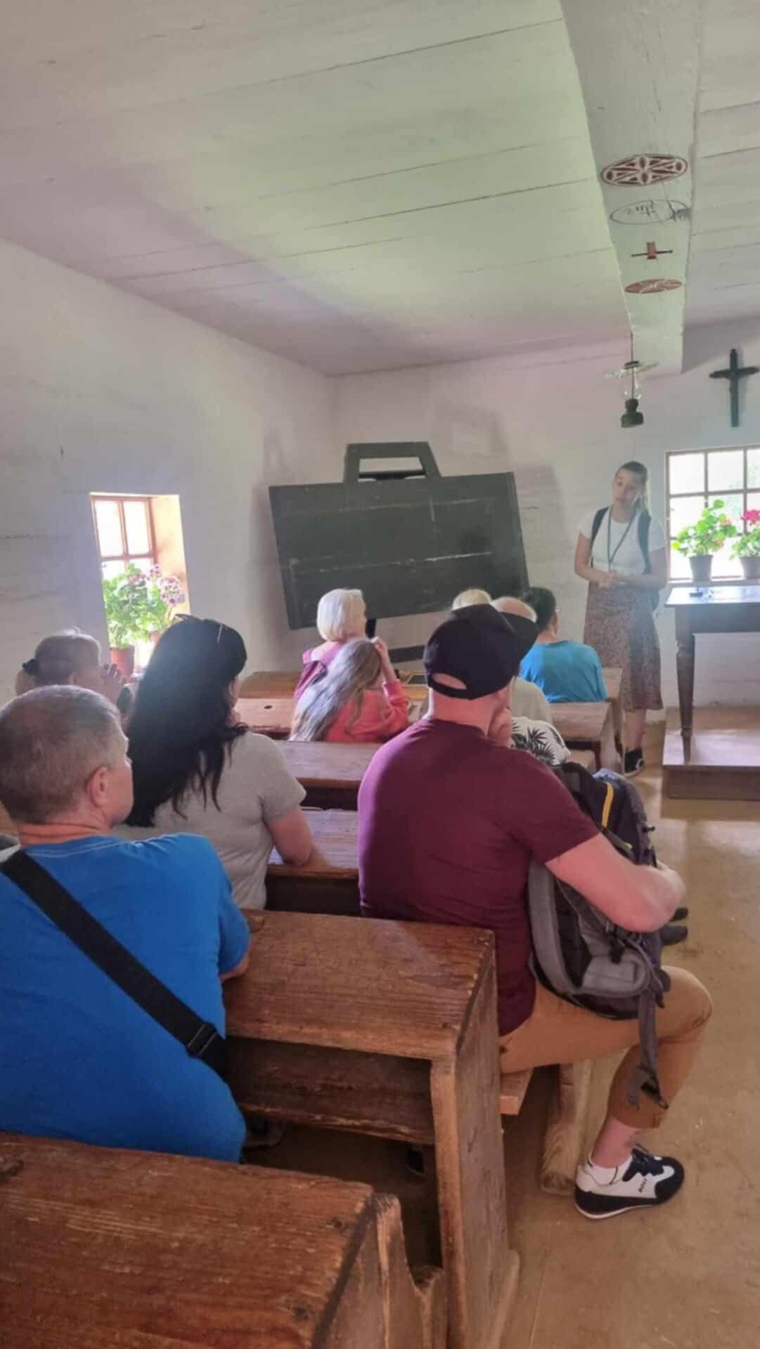 Grupa dorosłych siedzi w drewnianych ławkach naprzeciwko kobiety przemawiającej z przodu staromodnej klasy z tablicą do pisania kredą i krzyżem na ścianie.