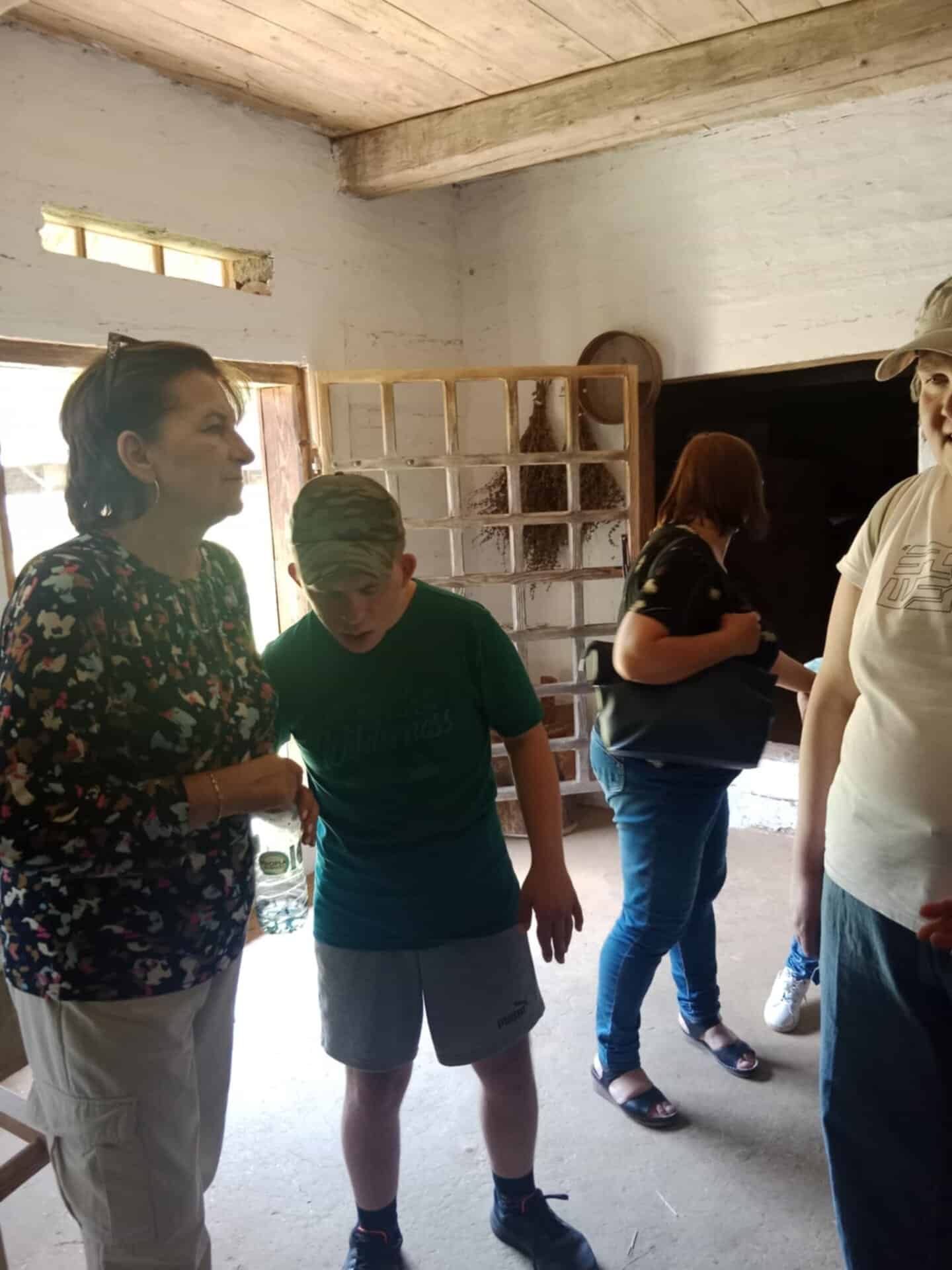 Cztery osoby stoją w rustykalnym pokoju z białymi ceglanymi ścianami i drewnianym sufitem, prowadząc rozmowę. Jedna osoba trzyma butelkę z wodą.