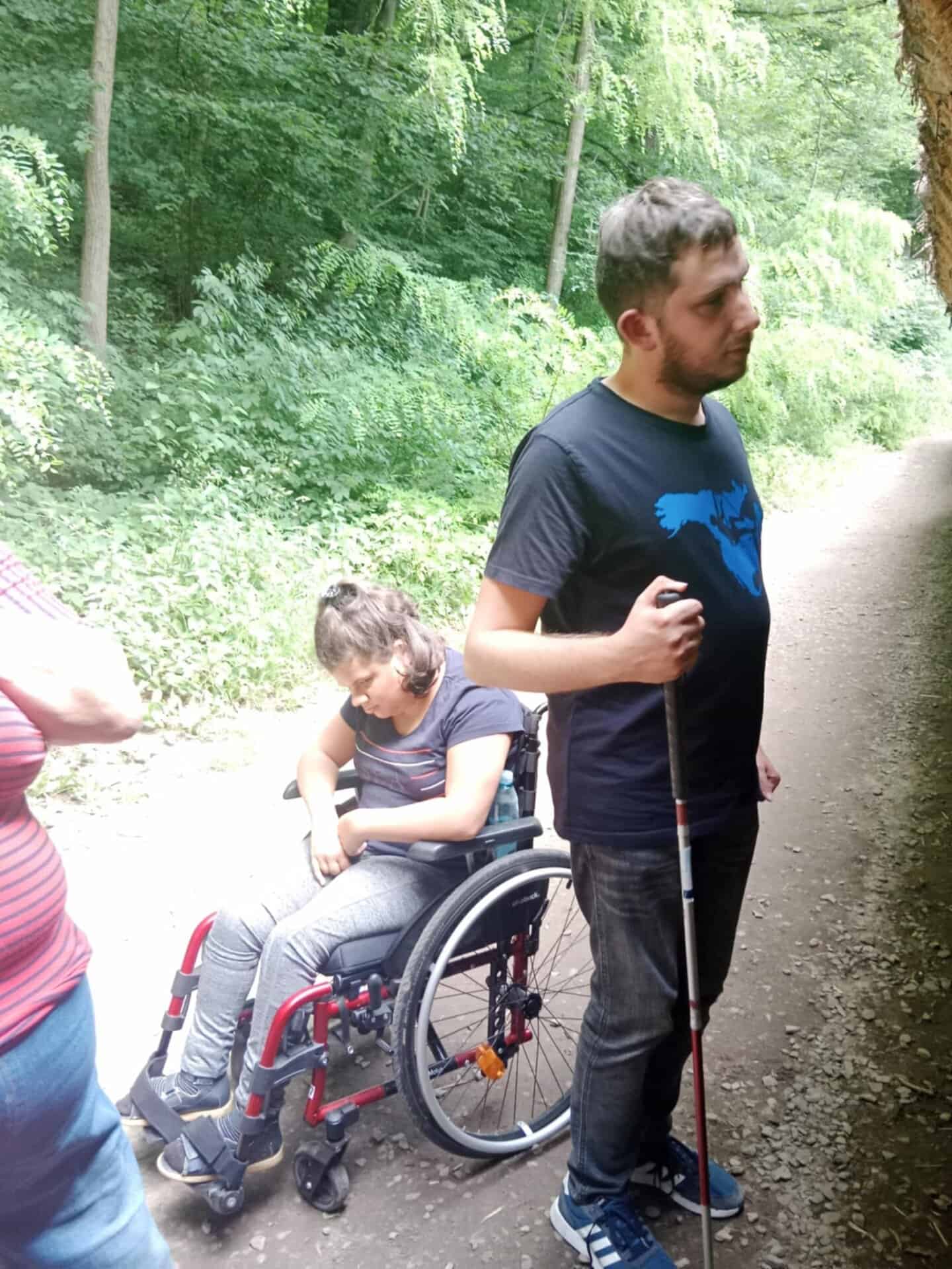 Mężczyzna trzymający białą laskę stoi na leśnej ścieżce obok młodej kobiety na wózku inwalidzkim; inna osoba jest częściowo widoczna po lewej stronie.